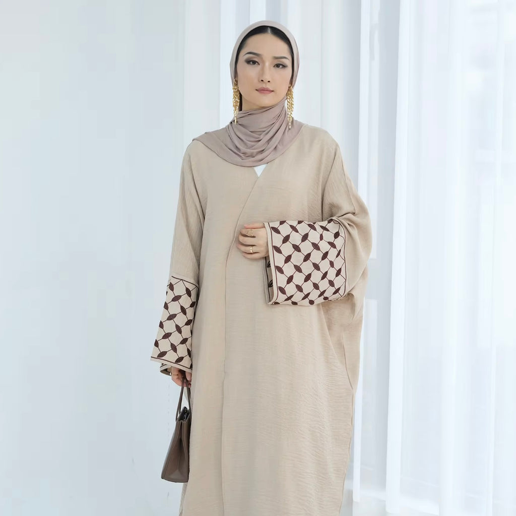 Woman wearing a beige abaya with patterned sleeves against a white curtain background