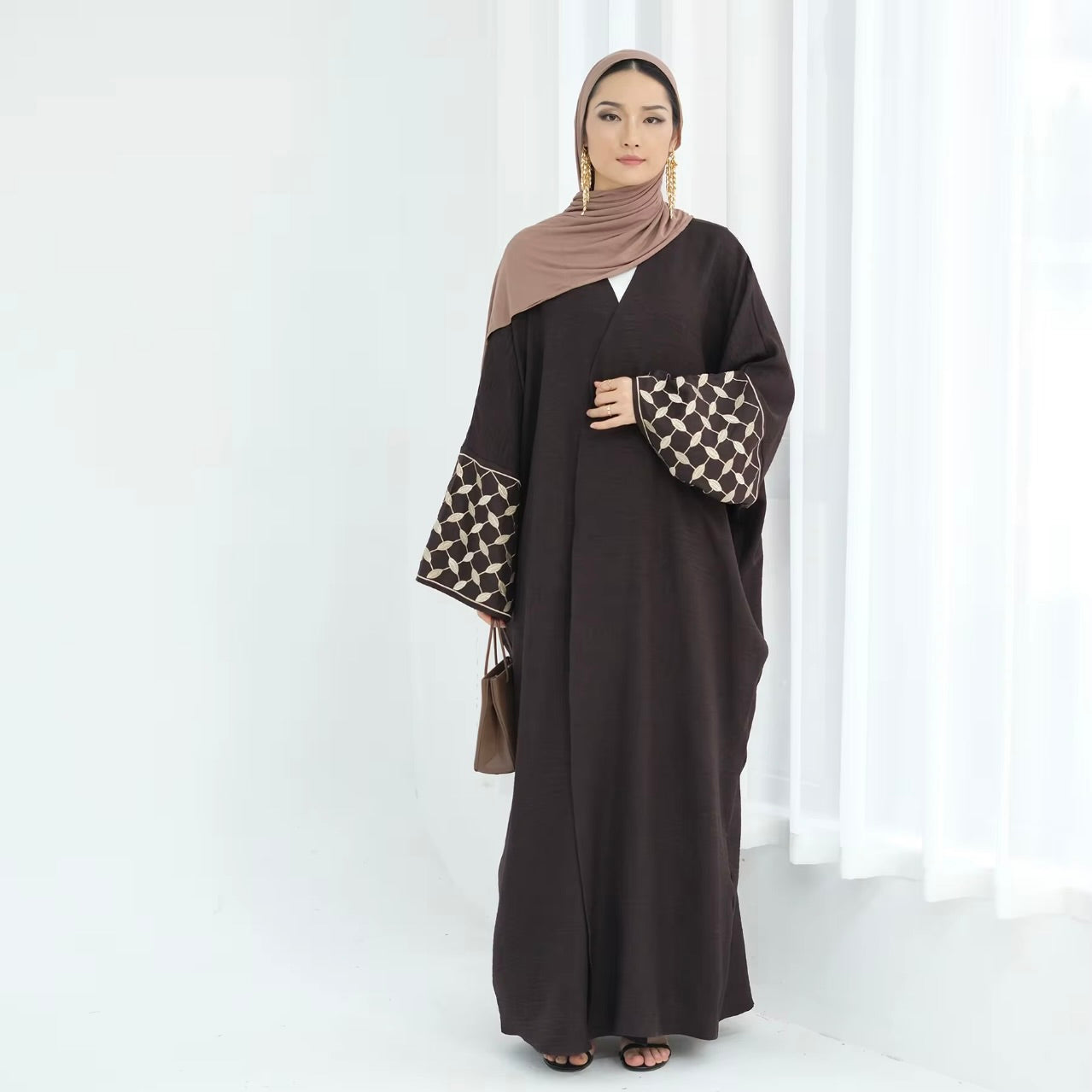 Woman wearing a dark brown abaya with patterned sleeves against a white curtain background
